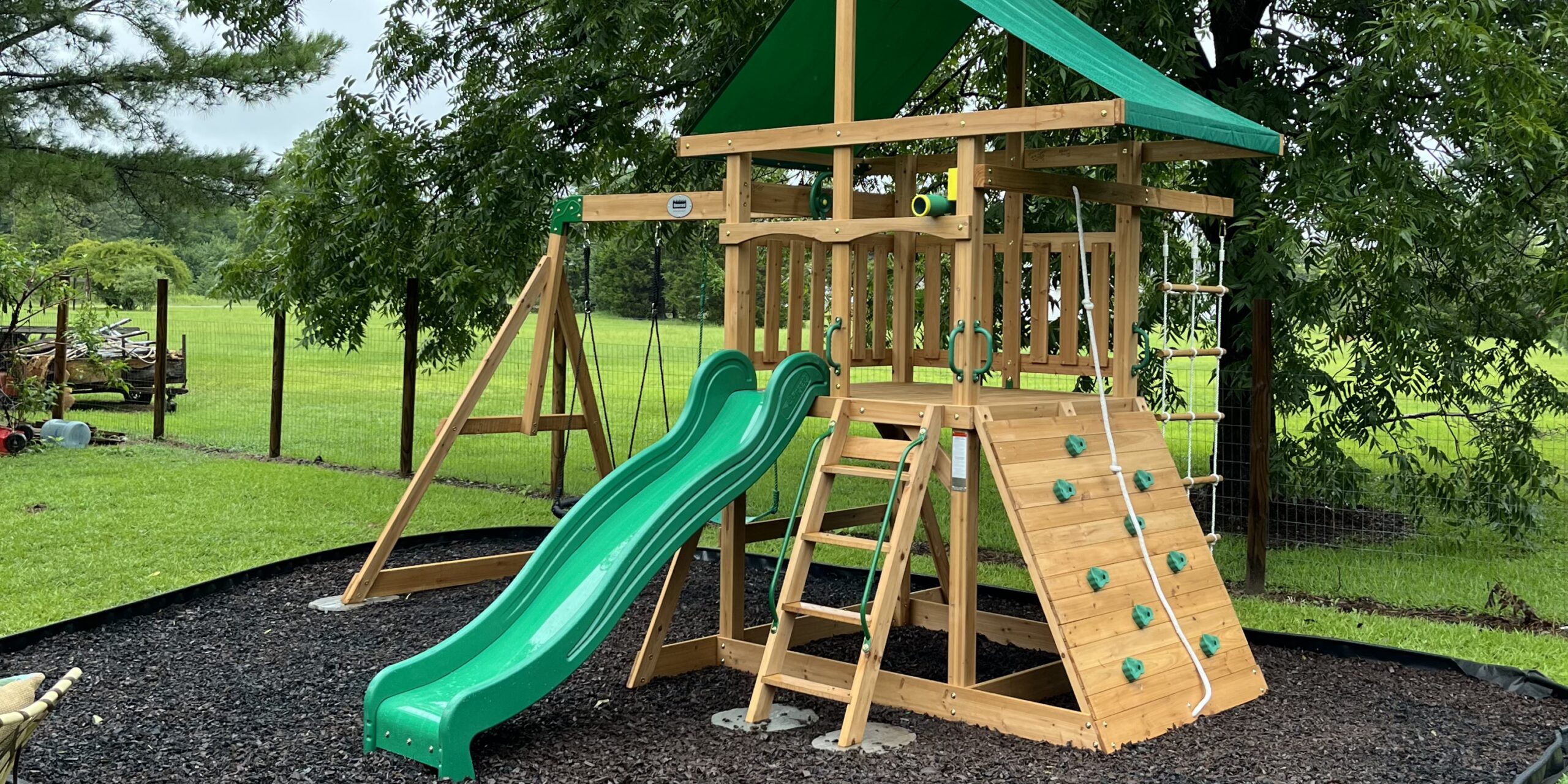 Wooden playset with slide, swing, and climbing wall installed on mulch surfacing in a backyard